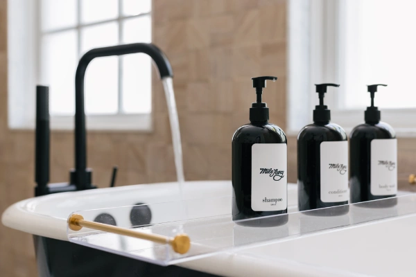 Close-up of soaking tub tray with branded shampoo and conditioner bottles beside matte black tub filler and wood-textured tile backdrop